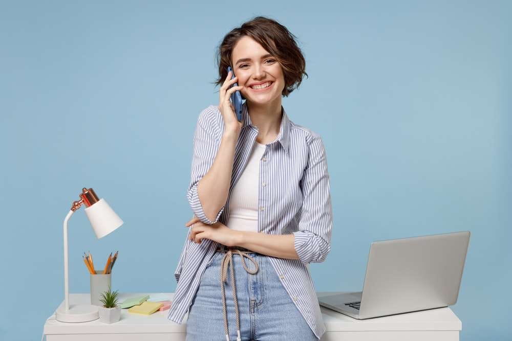 机とラップトップの横に立って電話で会話をしている若い女性。 机とラップトップの横に立って電話で会話をしている若い女性。
