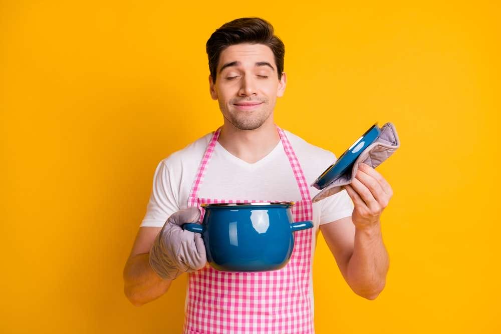 フランスで、エプロンと手袋をはめた男性が、黄色い背景を背景に、目を閉じて笑みを浮かべながら鍋と蓋を持っている。