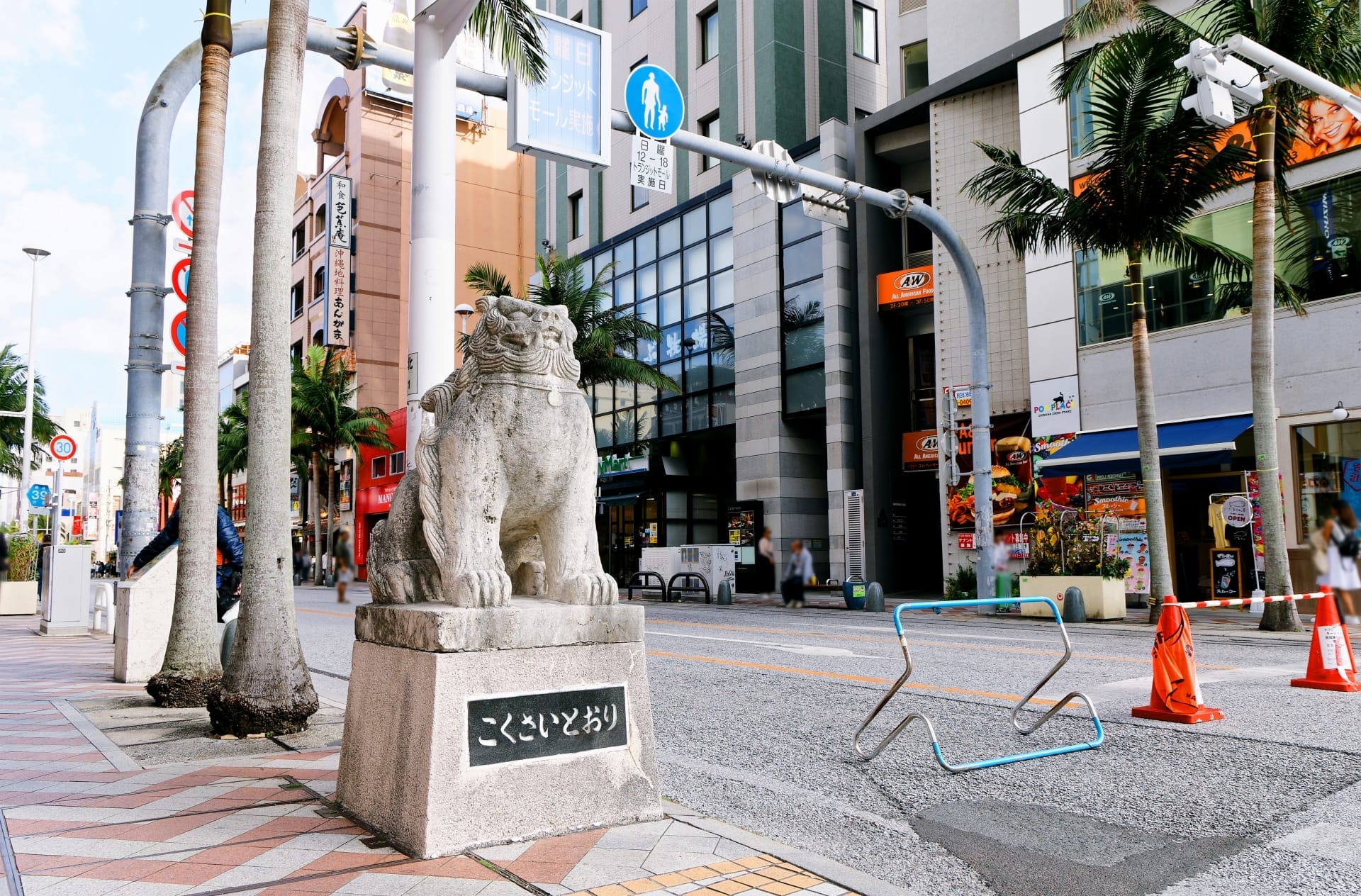 市街地の歩道にヤシの木や建物とともに佇む石造りのシーサー。近くのフランス語教室マンツーマン沖縄・那覇では、文化的魅力と教育の機会を融合させた、個人に合わせた語学クラスを提供しています。.