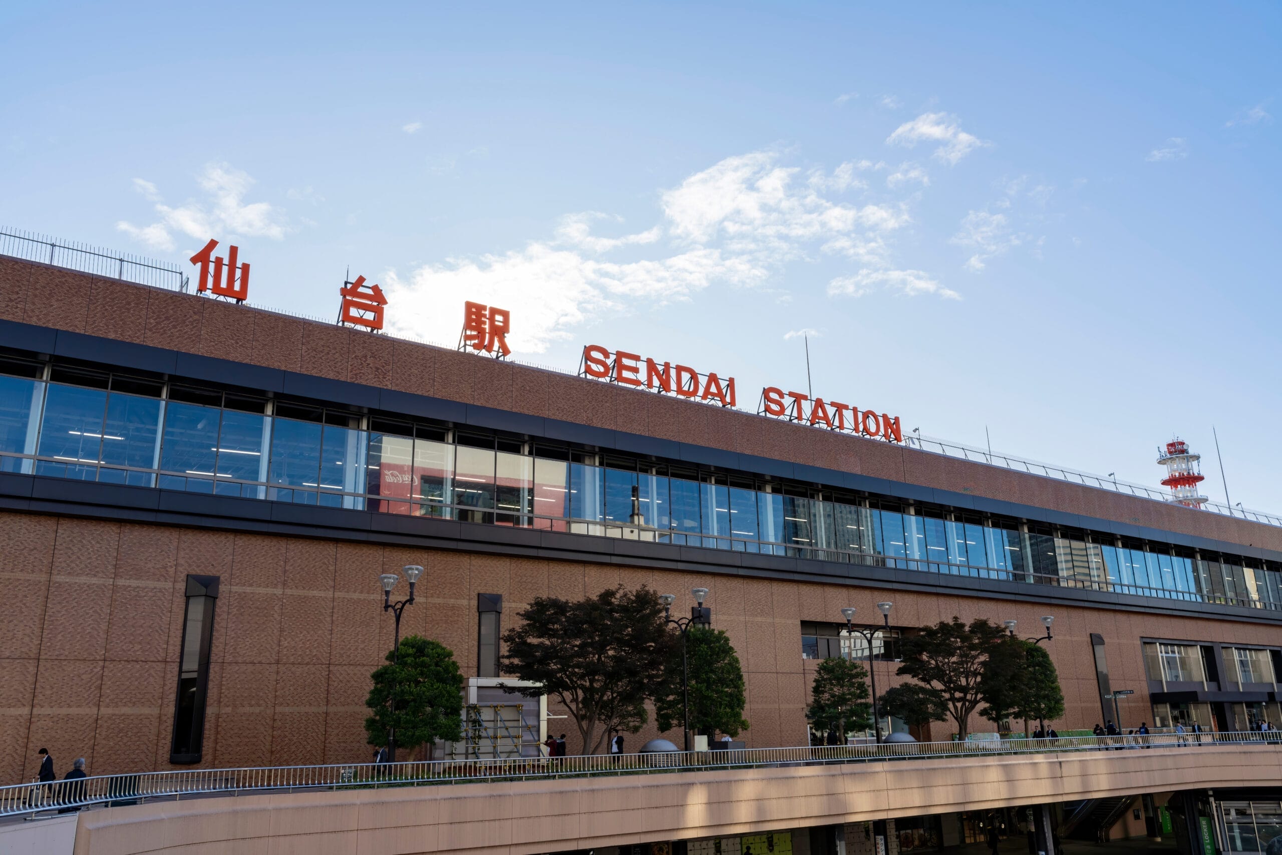 大きなレンガ造りの建物と、日本語と英語で書かれた目立つ標識が特徴的な仙台駅の外観。手前には澄んだ空の下の歩道と木々が広がり、宮城・仙台の活気あるエッセンスを捉えています。.